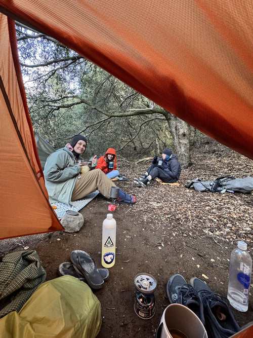 Backpackers sharing a meal under an orange tarp shelter with the stove in the foreground