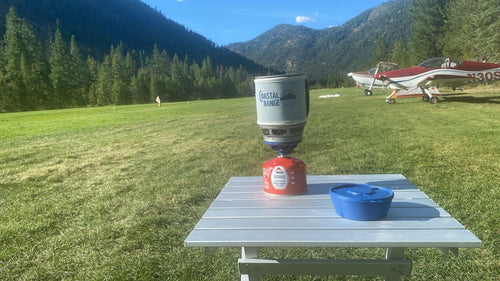 The Backpacker stove on a camp table at a grass airstrip with a bush plane parked behind