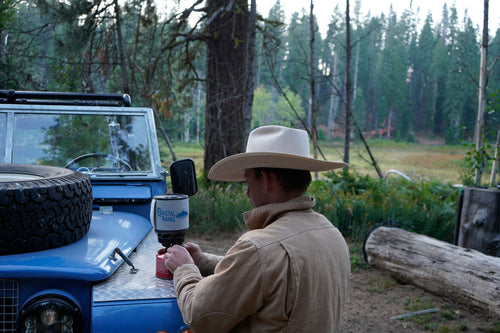 A person in a cowboy hat lighting the Backpacker stove on the fender of a blue Land Rover in the forest