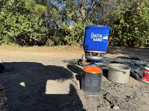 The Backpacker stove beside a separate metal pot and mug at a dusty campsite